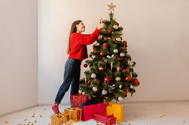 Mulher bonita sorridente e animada em um suéter vermelho em pé em casa decorando uma árvore de Natal cercada de presentes e caixas de presente