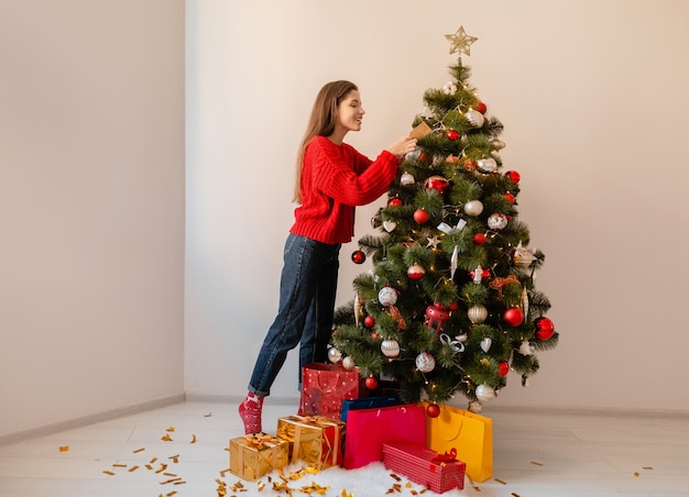 Mulher bonita sorridente e animada em um suéter vermelho em pé em casa decorando uma árvore de Natal cercada de presentes e caixas de presente