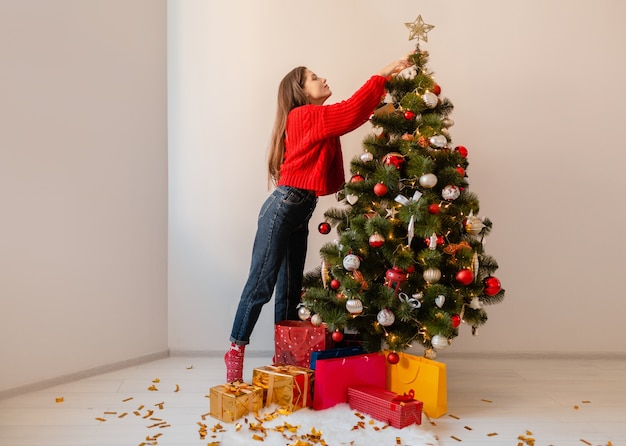 Mulher bonita sorridente e animada em um suéter vermelho em pé em casa decorando uma árvore de Natal cercada de presentes e caixas de presente