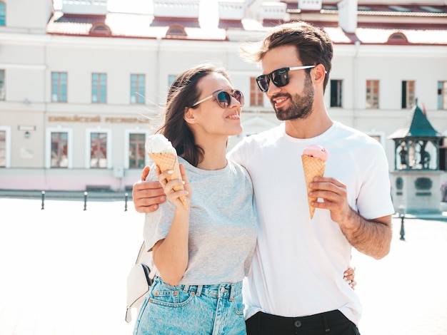 Foto grátis mulher bonita sorridente e seu namorado bonito mulher em roupas casuais de verão família alegre feliz casal posando no fundo da rua em óculos de sol comendo sorvete saboroso no cone de waffles