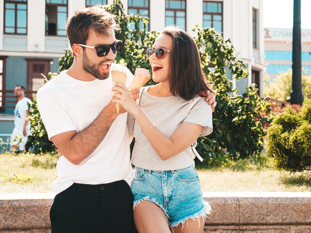 Mulher bonita sorridente e seu namorado bonito Mulher em roupas casuais de verão Família alegre feliz Casal posando no fundo da rua em óculos de sol Comendo sorvete saboroso no cone de waffles