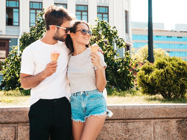 Mulher bonita sorridente e seu namorado bonito Mulher em roupas casuais de verão Família alegre feliz Casal posando no fundo da rua em óculos de sol Comendo sorvete saboroso no cone de waffles