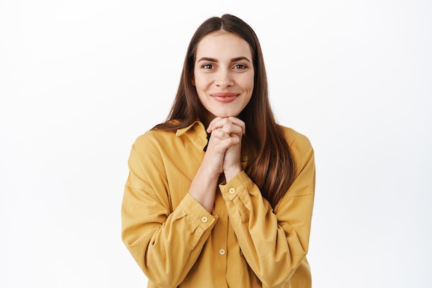 Mulher bonita sorridente esperando algo bom, esperando ou antecipando um presente, mãos dadas em um gesto esperançoso, em pé sobre uma parede branca