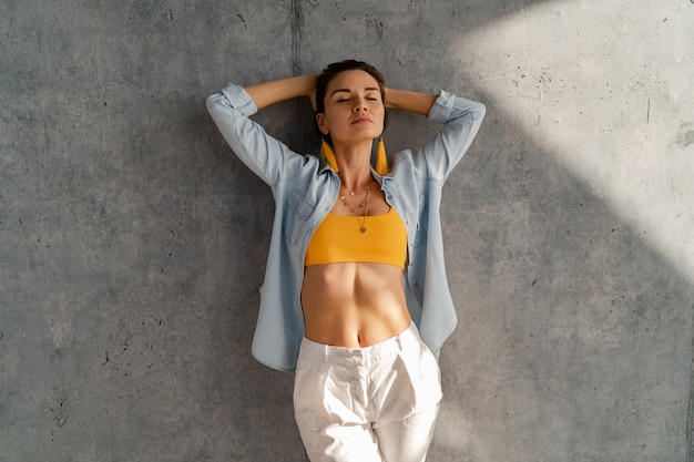 Mulher bonita sorridente feliz posando contra uma parede de concreto em um dia ensolarado, vestindo camisa jeans, blusa amarela e calça branca