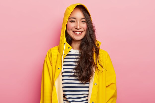 Mulher bonita sorridente gosta de vestir um macacão listrado quente, capa de chuva amarela com capuz, tem bom humor, sai com os amigos em dia de chuva