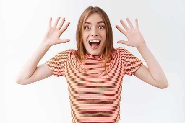 Mulher bonita, surpresa e entusiasmada em uma camiseta listrada, reage com emoção a notícias maravilhosas e surpreendentes, gritando atônita, sorrindo e levantando as mãos de alegria, em pé com fundo branco