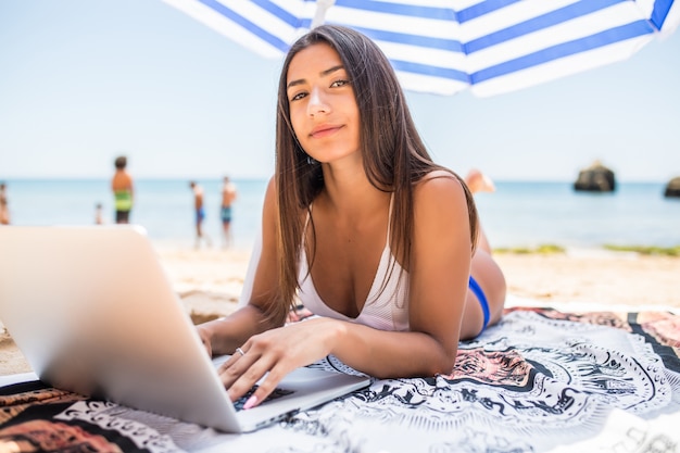 Mulher bonita trabalhando online no laptop enquanto estava deitado na praia sob o guarda-sol, perto do mar. garota freelancer sorridente feliz relaxando e usando o notebook para trabalho freelance de internet.