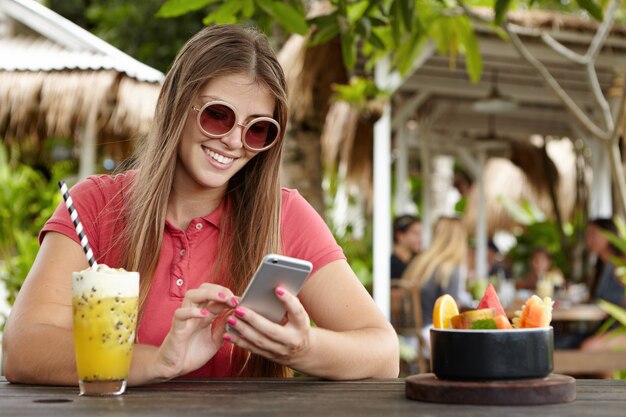 Mulher bonita vestindo camisa pólo e óculos de sol redondos, navegando na internet em seu telefone inteligente, desfrutando de comunicação online durante o almoço em um café na calçada