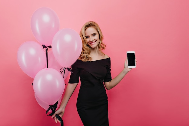 Mulher branca inspirada com penteado ondulado apresentando novo smartphone. Senhora caucasiana positiva segurando um monte de balões de festa rosa e sorrindo.