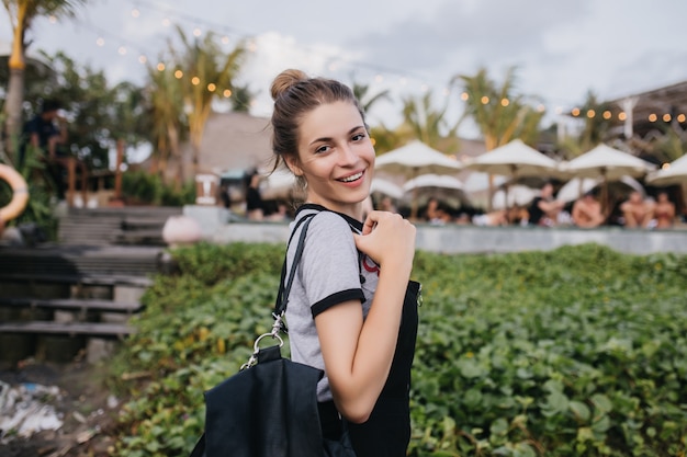 Mulher branca maravilhosa relaxando no resort de verão à noite. menina morena rindo com bolsa preta, olhando por cima do ombro na natureza.