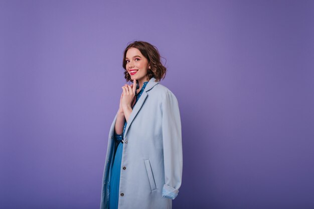 Mulher branca romântica com casaco elegante posando divertidamente na parede roxa. Foto interna de alegre modelo feminino encaracolado com corte de cabelo curto.