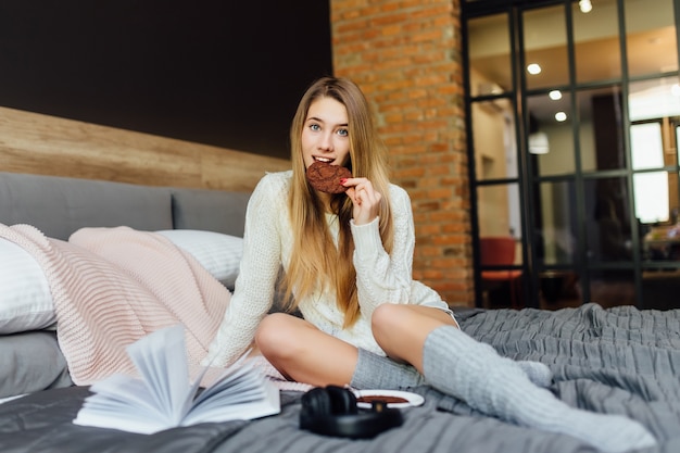 Mulher brincalhona relaxada vestindo camiseta, shorts e meias tendo preguiça de manhã, deitada na cama, tomando café da manhã