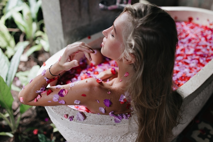 Foto grátis mulher bronzeada de cabelos compridos, desfrutando de spa no fim de semana. modelo feminino loiro deitado na banheira com pétalas de flores e arrepiando com os olhos fechados.