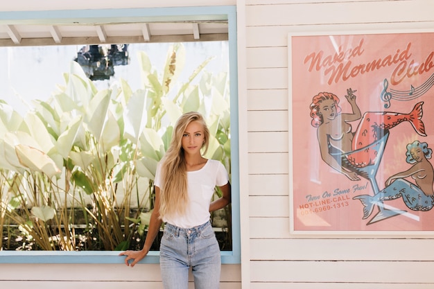 Foto grátis mulher bronzeada de cabelos compridos em pose confiante enquanto posava perto da janela. foto de uma garota entusiasmada em jeans e camiseta branca, sorrindo em dia de verão.