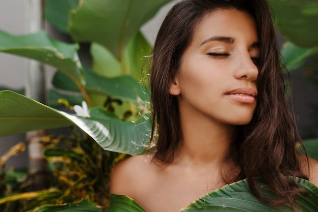 Mulher bronzeada sem maquiagem posando entre plantas tropicais com folhas enormes