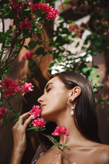 Mulher bronzeada sensual de biquíni, tocando o rosto com pétalas de flores tropicais, relaxando nas férias de verão.