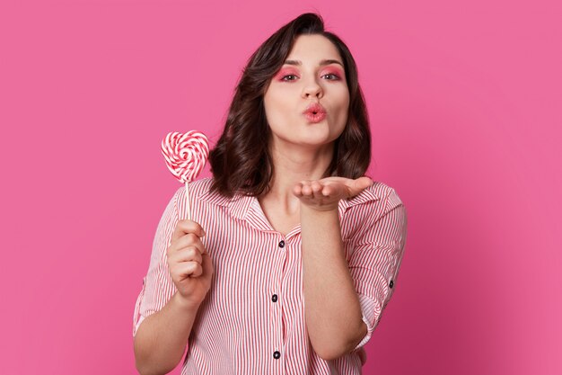 mulher carinhosa sopra beijo do ar, vestido com roupas da moda, detém doces, tem maquiagem brilhante, posa na rosada. Seu amor é por você. Adorável garota pin-up se diverte indoor.