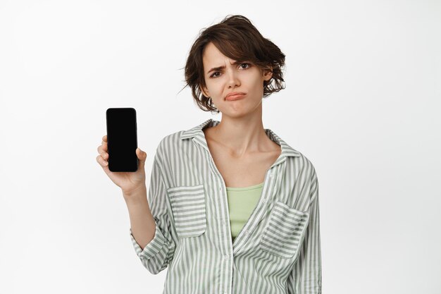 Mulher carrancuda decepcionada mostrando a tela do celular, de mau humor sobre algo no smartphone, reclamando do aplicativo, de pé sobre fundo branco