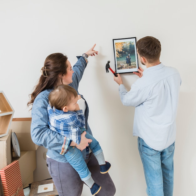 Mulher carregando seu filho dirigindo seu marido para consertar o quadro na parede