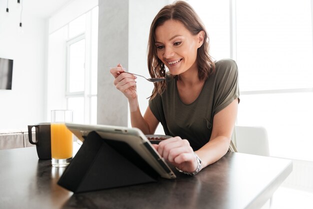 Mulher casual feliz comendo junto à mesa na cozinha e olhando para o computador tablet