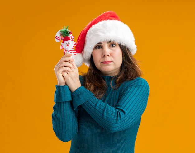 Mulher caucasiana adulta ansiosa com chapéu de Papai Noel segurando uma bengala de doce isolada em um fundo laranja com espaço de cópia