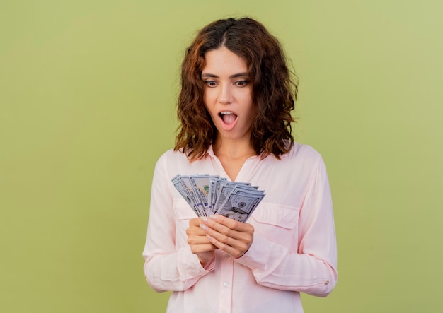 Foto grátis mulher caucasiana animada segurando e olhando para dinheiro isolado em um fundo verde com espaço de cópia