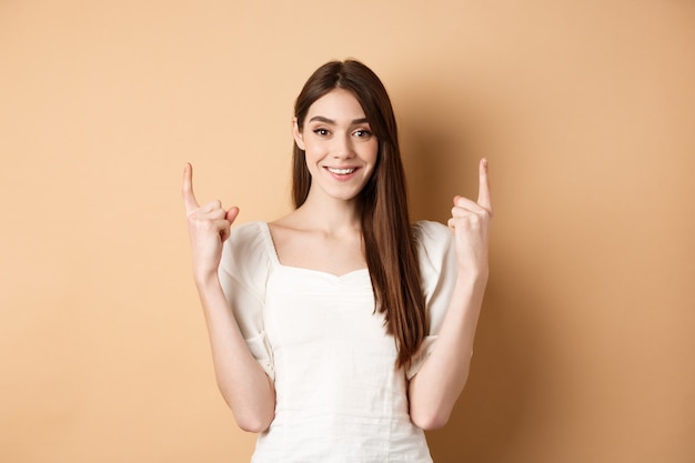 Mulher caucasiana atraente em vestido apontando dedos para cima, sorrindo e mostrando banner promo em pé.