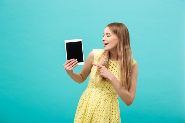 Mulher caucasiana atraente feliz apontando o dedo em um tablet para o espaço da cópia isolado sobre o fundo azul