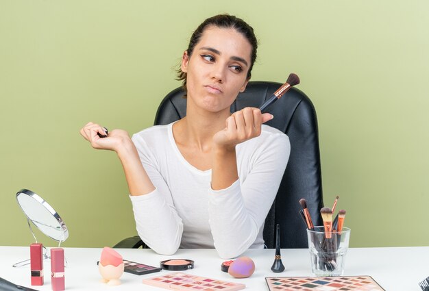 Mulher caucasiana bonita e descontente sentada à mesa com ferramentas de maquiagem segurando e olhando para o pincel de maquiagem