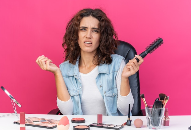 Mulher caucasiana bonita e descontente sentada à mesa com ferramentas de maquiagem segurando um pente