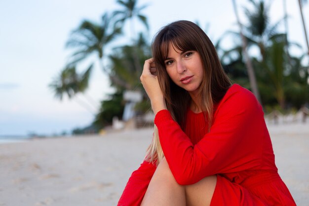 Mulher caucasiana com vestido vermelho de verão e clima romântico feliz na praia tropical de areia branca ao pôr do sol