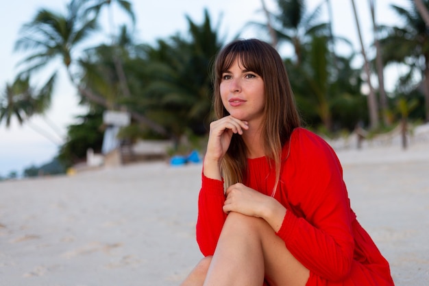 Mulher caucasiana com vestido vermelho de verão e clima romântico feliz na praia tropical de areia branca ao pôr do sol