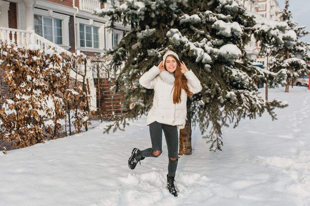 Mulher caucasiana dançando num lindo chapéu de malha. Retrato de corpo inteiro ao ar livre de uma senhora de cabelos compridos feliz em calças rasgadas brincando perto de uma árvore com neve.