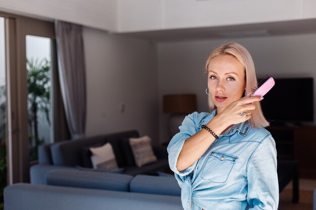 Mulher caucasiana de cabelos curtos, bonita, loira, com jaqueta jeans, escovando os cabelos com uma escova rosa em casa