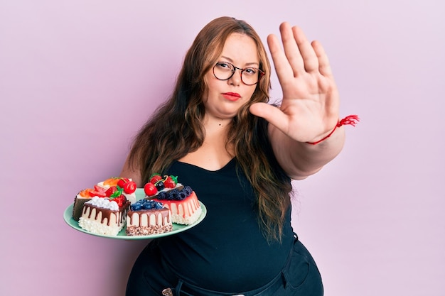 Foto grátis mulher caucasiana de tamanho grande segurando fatias de bolo com a mão aberta fazendo o sinal de stop com gesto de defesa de expressão séria e confiante