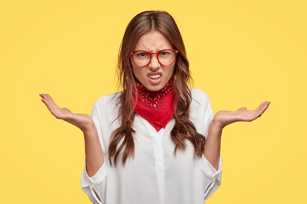 Mulher caucasiana descontente estende as mãos, franze a testa em desgosto, fica insatisfeita com algo desagradável, tem cabelos escuros penteados em tranças, fica encostada na parede amarela. isso é horrível!