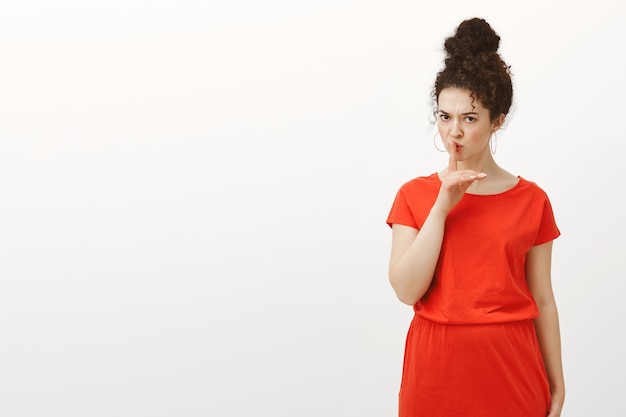 Mulher caucasiana estrita e bonita com cabelo encaracolado em um vestido vermelho casual, dizendo shh, fazendo um gesto de aviso de silêncio com o dedo indicador sobre a boca, franzindo a testa