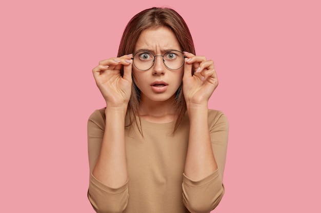 Foto grátis mulher caucasiana estupefata encara através dos óculos, não consegue acreditar nos próprios olhos, sendo emocional, estando em estupor com notícias repentinas, tem cabelo liso, vestida com roupas casuais, isolada sobre a parede rosa