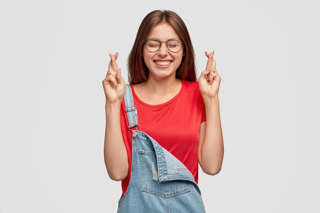 Mulher caucasiana feliz com cabelo escuro, mantém os dedos cruzados, reza por boa sorte, espera que os sonhos se tornem realidade tem um sorriso largo