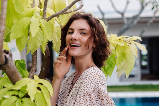 Mulher caucasiana feliz com vestido curto de verão do lado de fora do hotel villa perto da árvore e piscina azul