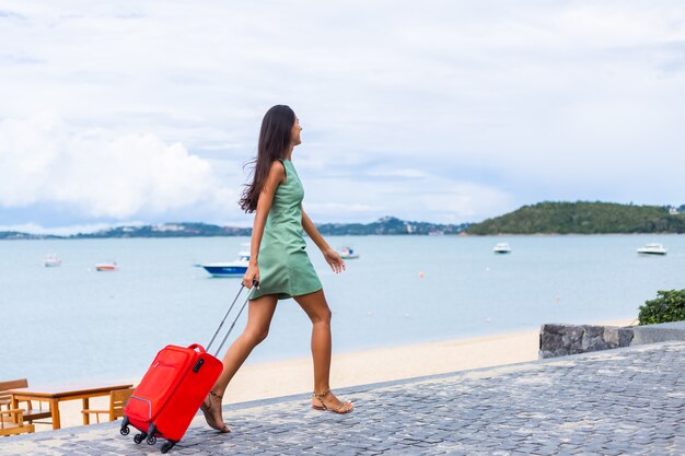 Mulher caucasiana feliz de cabelos longos e elegantes turistas em um vestido com mala vermelha fora do hotel