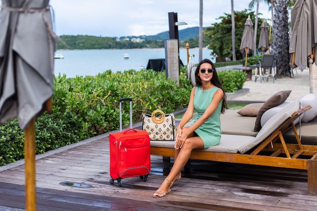 Mulher caucasiana feliz de cabelos longos e elegantes turistas em um vestido com mala vermelha fora do hotel