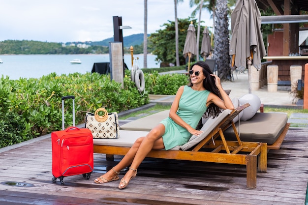 Mulher caucasiana feliz de cabelos longos e elegantes turistas em um vestido com mala vermelha fora do hotel