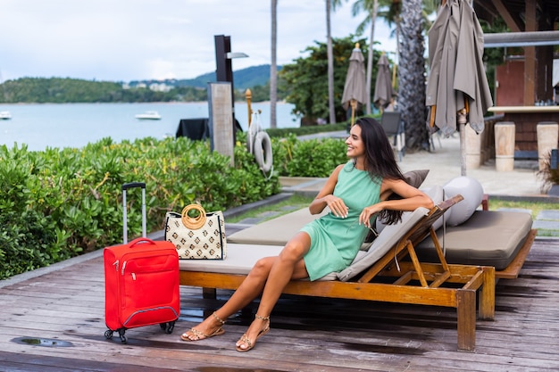 Mulher caucasiana feliz de cabelos longos e elegantes turistas em um vestido com mala vermelha fora do hotel