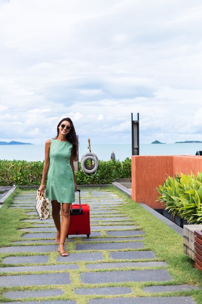 Mulher caucasiana feliz de cabelos longos e elegantes turistas em um vestido com mala vermelha fora do hotel