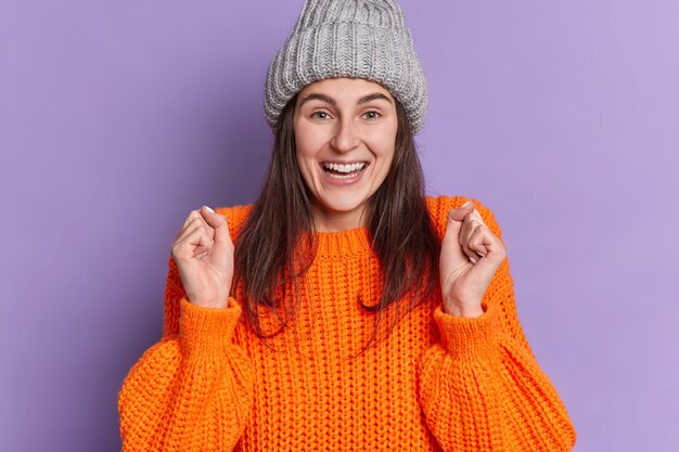 Mulher caucasiana morena positiva levanta os punhos cerrados sorrisos positivamente aguarda por algo muito bom acontecer usa um chapéu de camisola de malha.