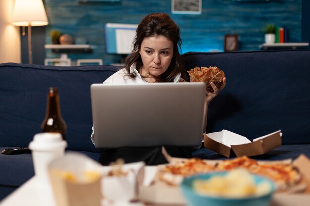 mulher caucasiana, sentada no sofá, comendo um hambúrguer delicioso e saboroso enquanto trabalhava no computador laptop