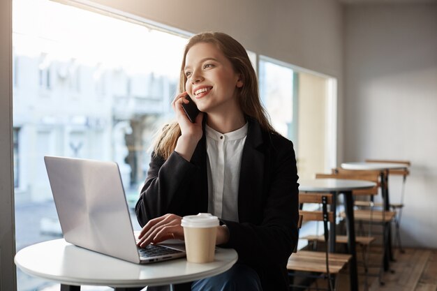 mulher caucasiana, sentado no café com laptop, tomando café, falando no smartphone