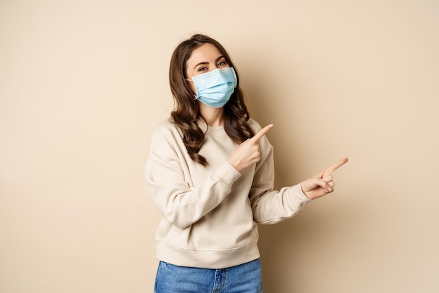 Mulher caucasiana sorridente feliz usando máscara facial médica da pandemia de coronavírus apontando os dedos r...