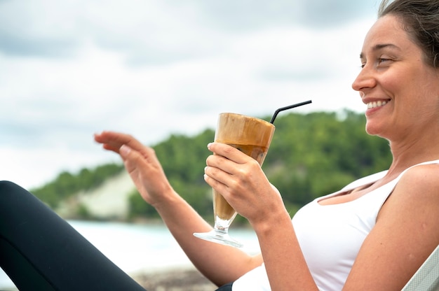 Mulher caucasiana sorridente segurando uma bebida de café em uma praia com espuma e canudo com colinas